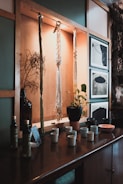 A wooden shelf displays hanging macramé art and a vase with dried branches. Below them, a collection of ceramic cups, a plant in a terracotta pot, bottles, and framed black and white photographs provide a cozy, rustic vibe.