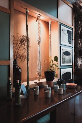 A wooden shelf displays hanging macramé art and a vase with dried branches. Below them, a collection of ceramic cups, a plant in a terracotta pot, bottles, and framed black and white photographs provide a cozy, rustic vibe.