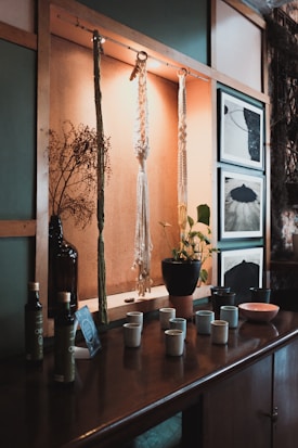 A wooden shelf displays hanging macramé art and a vase with dried branches. Below them, a collection of ceramic cups, a plant in a terracotta pot, bottles, and framed black and white photographs provide a cozy, rustic vibe.