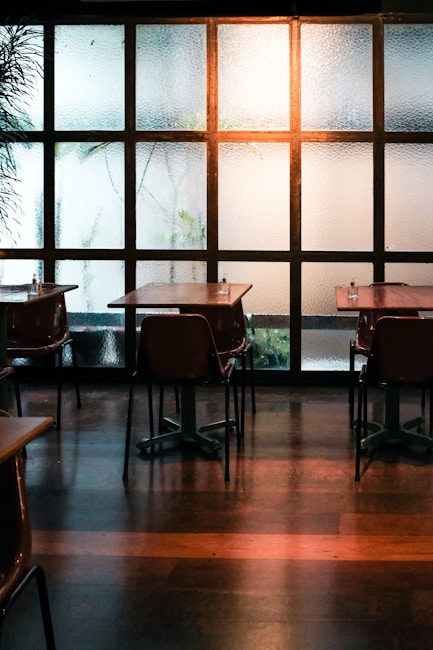 A quiet cafe interior with several empty tables and chairs arranged neatly. The room is dimly lit, with natural light filtering through frosted glass windows that have a grid-like pattern. There are some plants faintly visible outside, adding a hint of greenery.