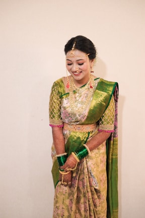 A woman is adorned in an elegant traditional sari featuring a combination of green and pink floral patterns. She wears elaborate gold jewelry, including a headpiece, necklace, earrings, and bangles. Her hands are decorated with henna, and she appears to be in a joyful or celebratory mood.