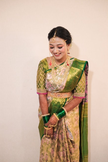 A woman is adorned in an elegant traditional sari featuring a combination of green and pink floral patterns. She wears elaborate gold jewelry, including a headpiece, necklace, earrings, and bangles. Her hands are decorated with henna, and she appears to be in a joyful or celebratory mood.