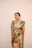 A smiling woman is dressed in traditional Indian attire, wearing a green and pink saree adorned with intricate patterns. She is accessorized with jewelry, including a necklace, earrings, and bangles. Henna designs cover her hands.