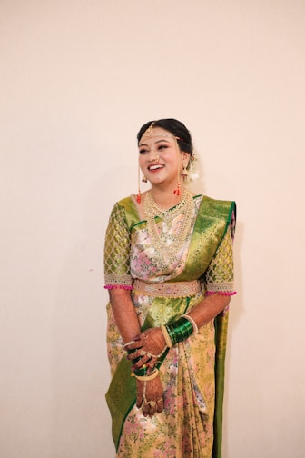 A smiling woman is dressed in traditional Indian attire, wearing a green and pink saree adorned with intricate patterns. She is accessorized with jewelry, including a necklace, earrings, and bangles. Henna designs cover her hands.