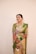 A smiling woman is dressed in traditional Indian attire, wearing a green and pink saree adorned with intricate patterns. She is accessorized with jewelry, including a necklace, earrings, and bangles. Henna designs cover her hands.