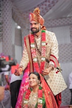A couple is dressed in traditional Indian wedding attire. The groom is placing a wedding necklace on the bride, with both wearing ornate outfits adorned with floral garlands. The groom wears a beige embroidered sherwani with a matching turban, while the bride is dressed in a green and red sari with heavy gold jewelry. The background is decorated with patterned drapes, and there are people standing around.