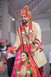 A couple is dressed in traditional Indian wedding attire. The groom is placing a wedding necklace on the bride, with both wearing ornate outfits adorned with floral garlands. The groom wears a beige embroidered sherwani with a matching turban, while the bride is dressed in a green and red sari with heavy gold jewelry. The background is decorated with patterned drapes, and there are people standing around.