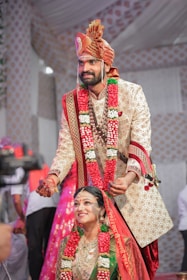 A couple is dressed in traditional Indian wedding attire. The groom is placing a wedding necklace on the bride, with both wearing ornate outfits adorned with floral garlands. The groom wears a beige embroidered sherwani with a matching turban, while the bride is dressed in a green and red sari with heavy gold jewelry. The background is decorated with patterned drapes, and there are people standing around.