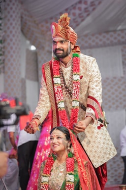 A couple is dressed in traditional Indian wedding attire. The groom is placing a wedding necklace on the bride, with both wearing ornate outfits adorned with floral garlands. The groom wears a beige embroidered sherwani with a matching turban, while the bride is dressed in a green and red sari with heavy gold jewelry. The background is decorated with patterned drapes, and there are people standing around.