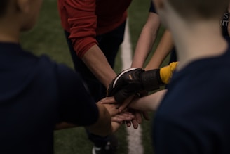 A group of people forming a huddle with their hands stacked together. The focus is on the hands, with arms wearing different clothing including a glove and a long-sleeved red shirt. The background shows a field with grass.