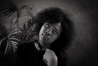 A black and white portrait of a woman with natural hair, positioned against a textured background with palm leaves. She is wearing a dark top and gazes directly at the camera with a calm expression, resting her chin on her hand.