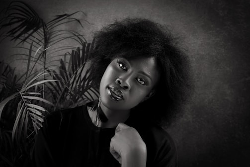 A black and white portrait of a woman with natural hair, positioned against a textured background with palm leaves. She is wearing a dark top and gazes directly at the camera with a calm expression, resting her chin on her hand.