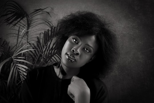 A black and white portrait of a woman with natural hair, positioned against a textured background with palm leaves. She is wearing a dark top and gazes directly at the camera with a calm expression, resting her chin on her hand.