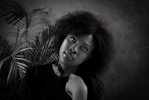 A black and white portrait of a woman with natural hair, positioned against a textured background with palm leaves. She is wearing a dark top and gazes directly at the camera with a calm expression, resting her chin on her hand.
