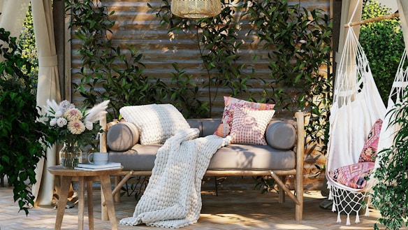 A cozy outdoor seating area featuring a wooden bench with plush cushions and a knitted throw. Next to the bench, a small round table holds a glass vase with a bouquet of flowers and a mug. On the right, a hanging hammock chair with patterned pillows adds to the relaxed atmosphere. The background consists of wooden paneling covered with lush green climbing plants, creating a tranquil, natural setting.