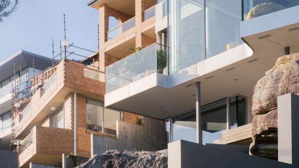 Modern residential buildings with large glass windows and brick facades are positioned on a rocky hill. The structures feature contemporary architectural elements such as balconies with glass railings. The use of natural rock and wood adds to the aesthetic.