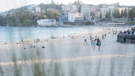 A beach scene with people enjoying various activities on the sand. The background includes buildings and trees, indicating an urban area nearby. The ocean is visible to the left, with calm waves approaching the shore. Volleyball nets are set up on the sand, and groups of people are scattered throughout, either resting or playing.