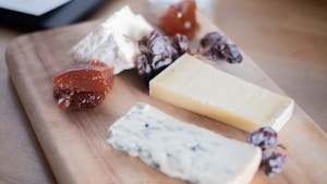 A wooden board displays various types of cheese, including a block of yellow cheese, a soft white cheese, and a blue cheese. Accompanying the cheese are several red jelly-like spreads and small clusters of dark dried fruit, possibly dates.
