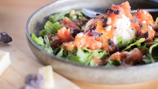 A close-up of a bowl filled with a fresh salad composed of leafy greens topped with a chunky, vibrant red tomato-based dressing. Visible are pieces of white cheese and scattered dark bits that could be olives or pepper flakes. A spoon rests in the bowl, ready for serving.