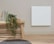 A minimalist interior features a light wooden bench against a beige wall. On the bench are a white and wooden watering can and a woven basket with a green leafy plant cascading down. Adjacent to the bench is a white square panel mounted on the wall. The floor is light wood and there is a grey textured rug nearby.
