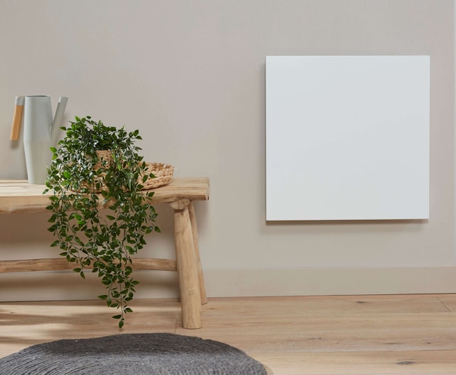 A minimalist interior features a light wooden bench against a beige wall. On the bench are a white and wooden watering can and a woven basket with a green leafy plant cascading down. Adjacent to the bench is a white square panel mounted on the wall. The floor is light wood and there is a grey textured rug nearby.