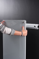 A person is holding a glass or acrylic panel with metal fixtures, likely installing or removing it from a wall mount bracket against a black background. The person is wearing a long-sleeved shirt with geometric patterns and has red nail polish.
