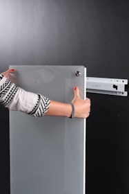 A person is holding a glass or acrylic panel with metal fixtures, likely installing or removing it from a wall mount bracket against a black background. The person is wearing a long-sleeved shirt with geometric patterns and has red nail polish.