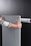 A person is holding a glass or acrylic panel with metal fixtures, likely installing or removing it from a wall mount bracket against a black background. The person is wearing a long-sleeved shirt with geometric patterns and has red nail polish.