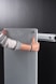 A person is holding a glass or acrylic panel with metal fixtures, likely installing or removing it from a wall mount bracket against a black background. The person is wearing a long-sleeved shirt with geometric patterns and has red nail polish.