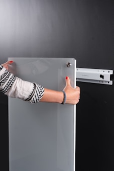 A person is holding a glass or acrylic panel with metal fixtures, likely installing or removing it from a wall mount bracket against a black background. The person is wearing a long-sleeved shirt with geometric patterns and has red nail polish.
