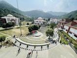 A scenic view of a college campus with a central amphitheater surrounded by greenery. The campus is set against a backdrop of lush, forested hills. Several dome-roofed buildings with red and white exteriors are visible. Students are walking and sitting around the area, while the sky is clear with some clouds.