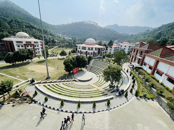 A scenic view of a college campus with a central amphitheater surrounded by greenery. The campus is set against a backdrop of lush, forested hills. Several dome-roofed buildings with red and white exteriors are visible. Students are walking and sitting around the area, while the sky is clear with some clouds.
