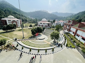 A scenic view of a college campus with a central amphitheater surrounded by greenery. The campus is set against a backdrop of lush, forested hills. Several dome-roofed buildings with red and white exteriors are visible. Students are walking and sitting around the area, while the sky is clear with some clouds.