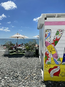 A pebbled beach scene with lounge chairs and umbrellas by the sea. The foreground features a colorful, decorated structure with artistic lettering and vibrant designs. People can be seen relaxing under parasols, enjoying the sunny weather. The sea meets a clear blue sky with scattered white clouds.