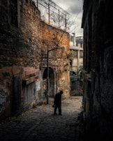 A narrow, cobblestone alleyway with dim lighting, bordered by aged stone and brick walls adorned with graffiti. A solitary figure in dark clothing is walking, head bowed, holding a small bag in one hand. Above the figure, a streetlamp provides sparse illumination. Tall buildings and a bare tree branch are visible in the background, with a cloudy sky overhead adding to the somber atmosphere.