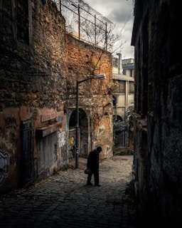 A narrow, cobblestone alleyway with dim lighting, bordered by aged stone and brick walls adorned with graffiti. A solitary figure in dark clothing is walking, head bowed, holding a small bag in one hand. Above the figure, a streetlamp provides sparse illumination. Tall buildings and a bare tree branch are visible in the background, with a cloudy sky overhead adding to the somber atmosphere.