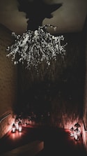 An intricately designed dark room features a ceiling installation resembling tree branches adorned with small, glowing lights. Below, a corner is softly illuminated by several lanterns casting a warm glow against textured walls.