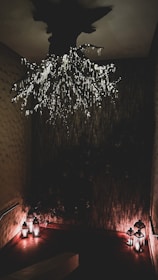 An intricately designed dark room features a ceiling installation resembling tree branches adorned with small, glowing lights. Below, a corner is softly illuminated by several lanterns casting a warm glow against textured walls.