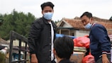 A group of people wearing masks are outdoors, likely transporting goods. One person in a black jacket stands near a metal railing while another individual in a blue jacket appears to be arranging or inspecting bags and boxes. The background features trees, rustic buildings with tiled roofs, and an overcast sky.