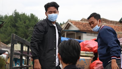 A group of people wearing masks are outdoors, likely transporting goods. One person in a black jacket stands near a metal railing while another individual in a blue jacket appears to be arranging or inspecting bags and boxes. The background features trees, rustic buildings with tiled roofs, and an overcast sky.