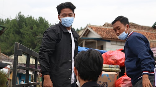 A group of people wearing masks are outdoors, likely transporting goods. One person in a black jacket stands near a metal railing while another individual in a blue jacket appears to be arranging or inspecting bags and boxes. The background features trees, rustic buildings with tiled roofs, and an overcast sky.