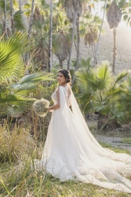 A bride stands in a lush garden setting surrounded by palm trees, wearing an elegant white wedding dress with a long train and holding a bouquet of flowers. Sunlight filters through the trees, creating a serene and picturesque atmosphere.