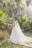 A bride stands in a lush garden setting surrounded by palm trees, wearing an elegant white wedding dress with a long train and holding a bouquet of flowers. Sunlight filters through the trees, creating a serene and picturesque atmosphere.