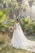 A bride stands in a lush garden setting surrounded by palm trees, wearing an elegant white wedding dress with a long train and holding a bouquet of flowers. Sunlight filters through the trees, creating a serene and picturesque atmosphere.
