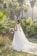 A bride stands in a lush garden setting surrounded by palm trees, wearing an elegant white wedding dress with a long train and holding a bouquet of flowers. Sunlight filters through the trees, creating a serene and picturesque atmosphere.