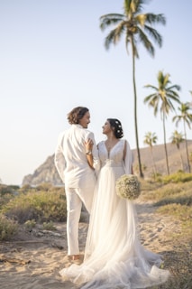A couple dressed in wedding attire walks along a sandy path surrounded by tropical greenery and tall palm trees under a clear sky. The woman wears a flowing white gown and holds a bouquet of flowers, while the man is dressed in a white suit. The scenery is serene and picturesque with distant rocky hills visible in the background.