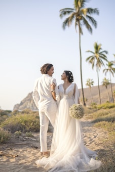 A couple dressed in wedding attire walks along a sandy path surrounded by tropical greenery and tall palm trees under a clear sky. The woman wears a flowing white gown and holds a bouquet of flowers, while the man is dressed in a white suit. The scenery is serene and picturesque with distant rocky hills visible in the background.