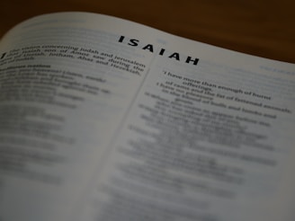 A close-up view of a printed page from a book, displaying text from the Book of Isaiah. The text is slightly out of focus, emphasizing the title 'ISAIAH' at the top. The page is angled, showing more text in the background.