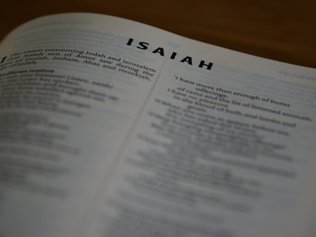A close-up view of a printed page from a book, displaying text from the Book of Isaiah. The text is slightly out of focus, emphasizing the title 'ISAIAH' at the top. The page is angled, showing more text in the background.