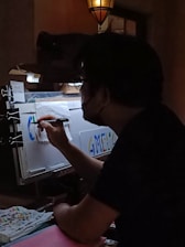 A person is seen working on a colorful piece of art in a dimly lit room. The artwork involves letters and vibrant colors, and is being created on a sheet held by clips. The environment suggests a creative workspace with a focus light and scattered art supplies.